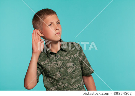 Close-up portrait of a blonde teenage boy in a green shirt with palm print posing against a blue studio background. Concept of sincere emotions. Close-up portrait of a blonde teenage boy in a green shirt with palm print posing against a blue studio background. Concept of sincere emotions. 89834502