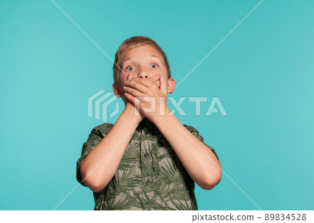 Close-up portrait of a blonde teenage boy in a green shirt with palm print posing against a blue studio background. Concept of sincere emotions. Close-up portrait of a blonde teenage boy in a green shirt with palm print posing against a blue studio background. Concept of sincere emotions. 89834528