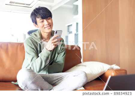 A young man cross-legged on the sofa and looking at his smartphone 89834923