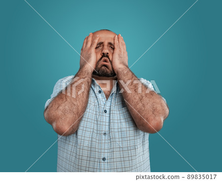 Close-up portrait of a brunet middle-aged man with beard, dressed in a light checkered shirt and posing against a blue background. 89835017