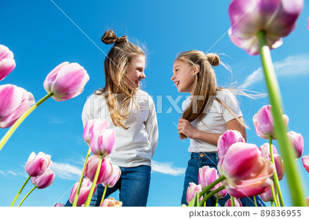 Two girls bend over blooming tulips against the blue sky, view from below. 89836955