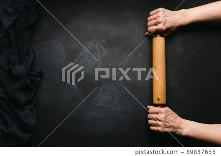 Two female hands hold a wooden rolling pin for rolling dough on a black table, top view. Place for an inscription 89837633