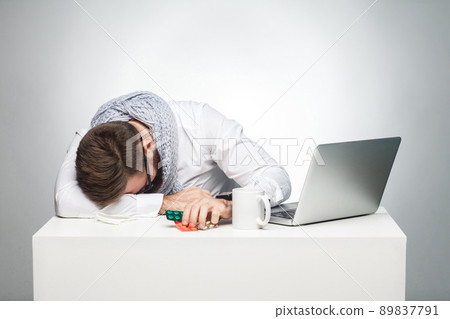 With temperature in the work station. Portrait of illness manager in white shirt are sitting in office is snoozing at his work place near laptop. Indoor, studio shot, gray background, isolated With temperature in the work station. Portrait of illness manager in white shirt are sitting in office is snoozing at his work place near laptop. Indoor, studio shot, gray background, isolated 89837791