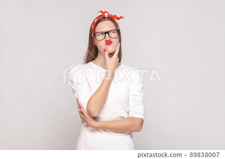 portrait of beautiful emotional young woman in white t-shirt with freckles, black glasses, red lips and head band touching her chin and thinking. indoor studio shot, isolated on light gray background. portrait of beautiful emotional young woman in white t-shirt with freckles, black glasses, red lips and head band touching her chin and thinking. indoor studio shot, isolated on light gray background. 89838007