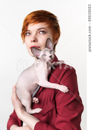 Canadian Sphynx Cat sitting on shoulder of hipster redhead young woman. Selective focus on foreground domestic hairless kitten, shallow depth of field. Studio shot, white background. Part of series 89838083