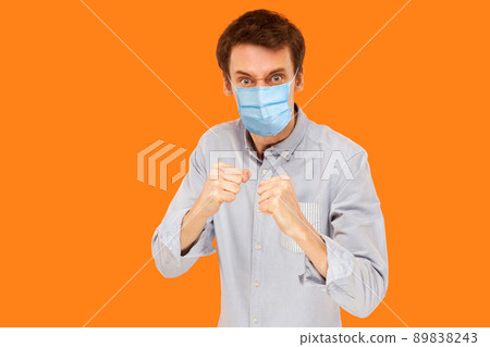 Portrait of angry young worker man with surgical medical mask standing in boxing fists and looking at camera and ready to attack against virus. indoor studio shot isolated on orange background. Portrait of angry young worker man with surgical medical mask standing in boxing fists and looking at camera and ready to attack against virus. indoor studio shot isolated on orange background. 89838243
