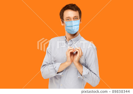 I love you. Portrait of joyful happy young worker man with surgical medical mask standing with heart love gesture and looking at camera with smile. indoor studio shot isolated on orange background. 89838344