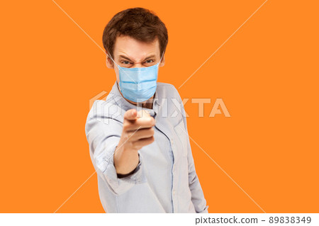 Hey you. Portrait of angry young worker man with surgical medical mask standing pointing and scolding at camera with mad face. indoor studio shot isolated on orange background. Hey you. Portrait of angry young worker man with surgical medical mask standing pointing and scolding at camera with mad face. indoor studio shot isolated on orange background. 89838349