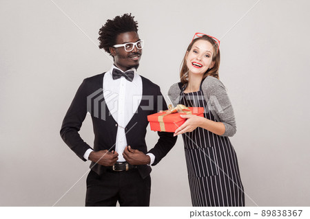 Freckled woman holding gift box, african man standing near and smiling, it was he who gave her a gift. Studio shot, gray background 89838367