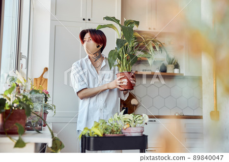 Young Asian guy in mask holding flower pot with green plant on the kitchen 89840047