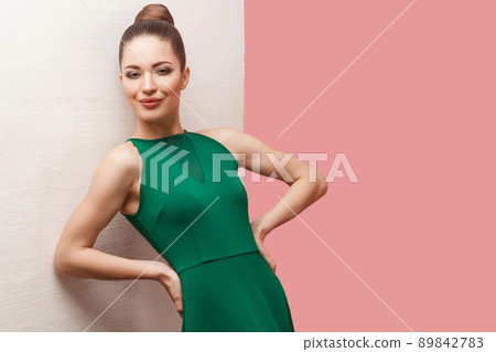 Portrait of happy beautiful young woman with bun hairstyle and makeup in green dress standing with hands on waist and looking at camera, lean on white wall. studio shot, isolated on pink background. 89842783