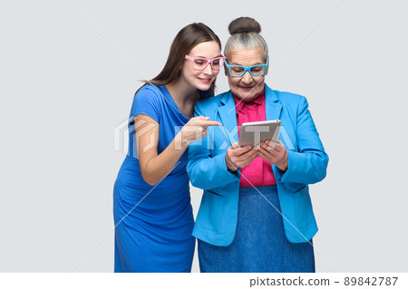 Young daughter in blue dress showing, pointing finger at tablet display in hands of aged woman. family relations between granddaughter and grandmother. indoor, studio shot, isolated on gray background 89842787