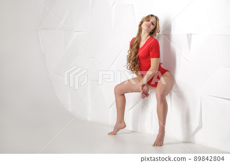 beautiful sexy girl in red bodysuit with long curly blonde hairstyle and makeup and perfect body lean and posing on white wall and looking at camera. good emotions and feelings, indoor studio shot. 89842804