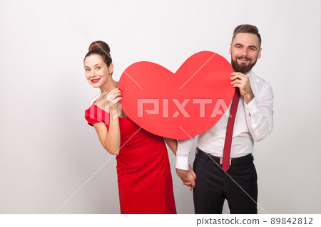 Attractive couple toothy smile, look out from behind big heart. Indoor, studio shot, isolated on gray background Attractive couple toothy smile, look out from behind big heart. Indoor, studio shot, isolated on gray background 89842812