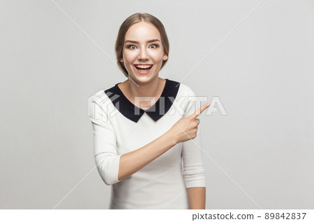 Businesswoman pointing finger at copy space, toothy smiling and looking at camera with amazed face. Studio shot, gray background 89842837