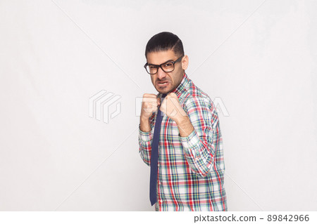 Angry bearded businessman in colorful checkered shirt, blue tie and black eyeglasses standing and looking at camera with aggressive face and fist. indoor studio shot, isolated on light grey background 89842966
