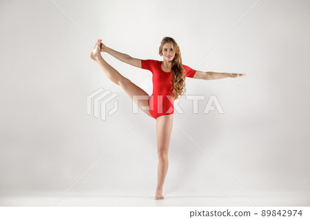 Happy blonde woman in red leotard standing on one leg and doing tree eka pada jathara parivartanasana yoga pose, indoor studio shot on light gray background. healthy lifestyle and leisure activity. Happy blonde woman in red leotard standing on one leg and doing tree eka pada jathara parivartanasana yoga pose, indoor studio shot on light gray background. healthy lifestyle and leisure activity. 89842974