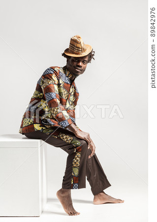 Happy fashion african man in traditional clothes sit on cube . Indoor, isolated on gray background Happy fashion african man in traditional clothes sit on cube . Indoor, isolated on gray background 89842976