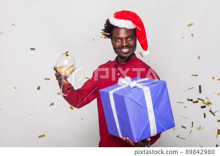 Around happiness afro man in red cap, flies gold metaphane, man holding champagne glass and gift box, looking at camera and toothy smiling. Studio shot. Gray background Around happiness afro man in red cap, flies gold metaphane, man holding champagne glass and gift box, looking at camera and toothy smiling. Studio shot. Gray background 89842980