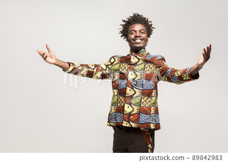 Friendly afro man in traditional clothes toothy smile, say welcome. Indoor, isolated on gray background 89842983