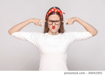 i dont want to hear you. portrait of unsatisfied emotional young woman in white t-shirt with freckles, black glasses, red lips and head band. indoor studio shot, isolated on light gray background. i dont want to hear you. portrait of unsatisfied emotional young woman in white t-shirt with freckles, black glasses, red lips and head band. indoor studio shot, isolated on light gray background. 89842991