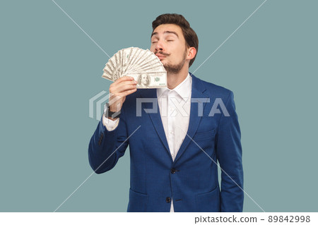 Handsome businessman in blue jacket standing and holding many dollars in hand and smelling and feeling smell of richness with closed eyes. Indoor, studio shot isolated on light blue background. 89842998