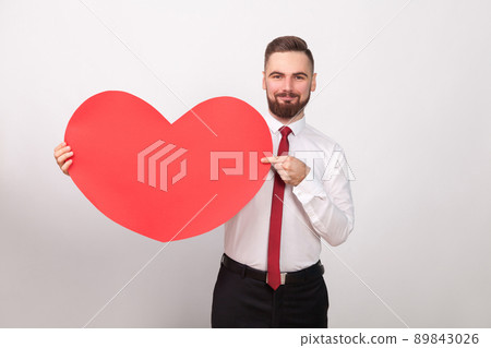 Perfect businessman smiling, pointing finger at big red heart. Indoor, studio shot, isolated on gray background 89843026