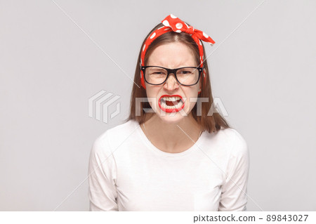 angry face screaming portrait of anger crazy bossy emotional young woman in white t-shirt with freckles, black glasses, red lips and head band. indoor studio shot, isolated on light gray background. angry face screaming portrait of anger crazy bossy emotional young woman in white t-shirt with freckles, black glasses, red lips and head band. indoor studio shot, isolated on light gray background. 89843027