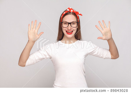 hey, nice to see you. portrait of beautiful emotional young woman in white t-shirt with freckles, black glasses, red lips and head band. indoor studio shot, isolated on light gray background. 89843028
