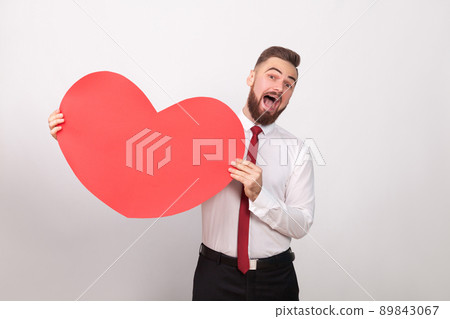 Funny businessman toothy smiling, holding big red heart. Indoor, studio shot, isolated on gray background 89843067