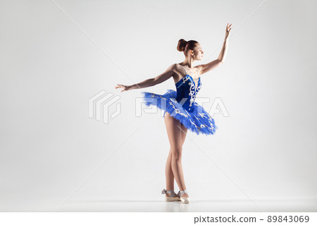 attractive ballerina with bun collected hair wearing blue dress and pointe shoes performing in white studio. indoor, studio shot. 89843069