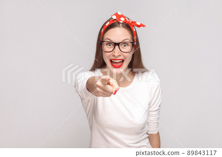 its it you? pointing finger at camera. portrait of beautiful emotional young woman in white t-shirt with freckles, black glasses, red lips and head band. indoor, isolated on light gray background. 89843117