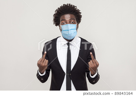 Go away from here Portrait of angry young man wearing black suit with medical mask standing and looking with aggressive face and showing middle finger. indoor studio shot isolated on gray background. 89843164