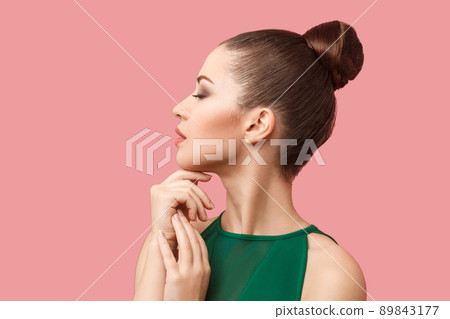 Profile side view portrait of calm serious beautiful young woman with bun hairstyle and makeup in green dress standing with closed eyes and touching her chin. studio shot, isolated on pink background. 89843177