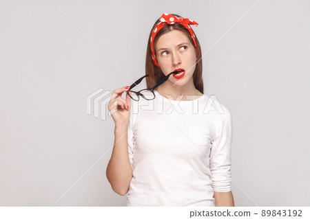Thoughtful portrait of beautiful emotional young woman in white t-shirt with freckles, black glasses in her mouth, red lips and head band. indoor studio shot, isolated on light gray background. 89843192