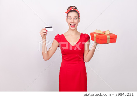 Shocked beautiful wonder woman, holding credit card, gift box.. Indoor, studio shot, isolated on gray background Shocked beautiful wonder woman, holding credit card, gift box.. Indoor, studio shot, isolated on gray background 89843231