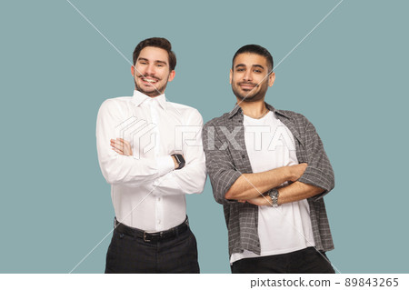 Portrait of two handsome satisfied bearded friends or partner standing with crossed hands and looking at camera with toothy smile. indoor studio shot, isolated on light blue background. Portrait of two handsome satisfied bearded friends or partner standing with crossed hands and looking at camera with toothy smile. indoor studio shot, isolated on light blue background. 89843265