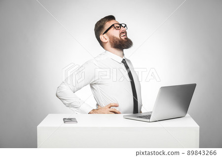 Side view of portrait of unhealphy upset young manager in white shirt and black tie are sitting in office and have strong backache or kidney pain. indoor, studio shot, isolated, gray background Side view of portrait of unhealphy upset young manager in white shirt and black tie are sitting in office and have strong backache or kidney pain. indoor, studio shot, isolated, gray background 89843266