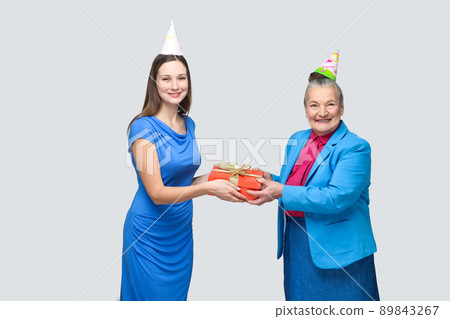 Grandmother in blue suit and hat holding red gift box and giving to her lovely granddaughter on her birthday. family happiness life event celebration. indoor studio shot, isolated on gray background Grandmother in blue suit and hat holding red gift box and giving to her lovely granddaughter on her birthday. family happiness life event celebration. indoor studio shot, isolated on gray background 89843267