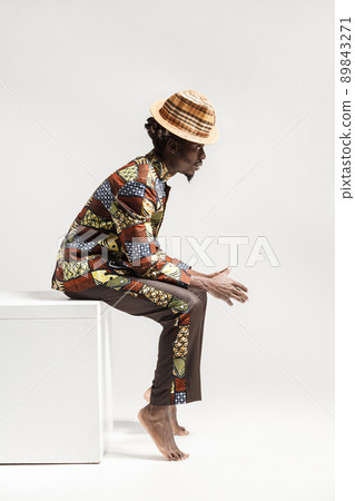 Unhappy afro man in traditional national clothes sit on cube. Indoor, isolated on gray background Unhappy afro man in traditional national clothes sit on cube. Indoor, isolated on gray background 89843271