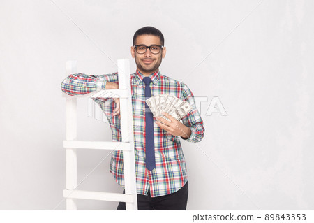 Portrait of successful rich handsome bearded young man in colorful checkered shirt with blue tie standing, holding fan of cash and lean on white stairs. Indoor studio shot, isolated on grey background Portrait of successful rich handsome bearded young man in colorful checkered shirt with blue tie standing, holding fan of cash and lean on white stairs. Indoor studio shot, isolated on grey background 89843353