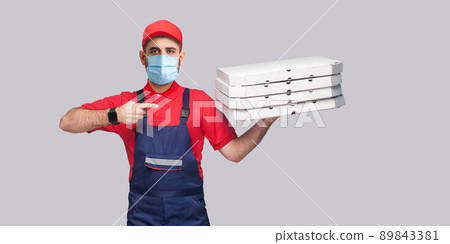 Delivery pizza on quarantine. Young man with surgical medical mask in blue uniform and red t-shirt standing, holding and pointing finger to stack of cardboard pizza boxes on grey background. Delivery pizza on quarantine. Young man with surgical medical mask in blue uniform and red t-shirt standing, holding and pointing finger to stack of cardboard pizza boxes on grey background. 89843381