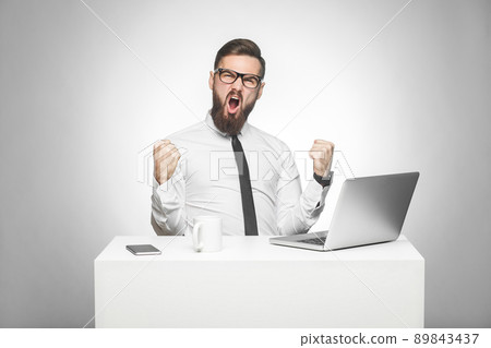 Portrait of handsome amazed satisfied positive bearded young businessman in white shirt and black tie are sitting in office and watching online football match with open mouth and triumphing. Indoor Portrait of handsome amazed satisfied positive bearded young businessman in white shirt and black tie are sitting in office and watching online football match with open mouth and triumphing. Indoor 89843437