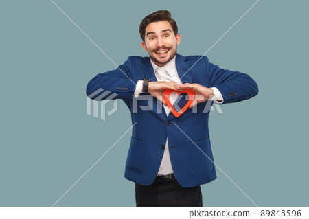 Handsome man in blue jacket and white shirt, standing and holding red heart shape and looking at camera with toothy smile. Indoor, studio shot isolated on light blue background 89843596
