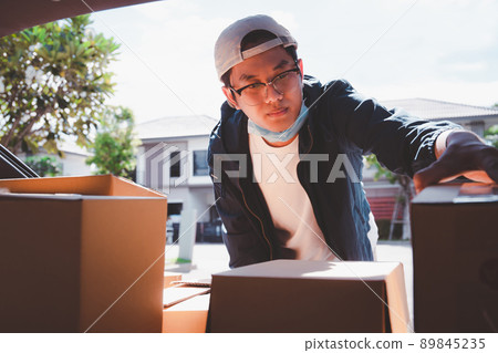 Asian postman with a hygiene protective face mask picking up a parcel package or parcel box from the truck for deliver to customer's door. New Normal concept during covid-19 or coronavirus pandemic Asian postman with a hygiene protective face mask picking up a parcel package or parcel box from the truck for deliver to customer's door. New Normal concept during covid-19 or coronavirus pandemic 89845235