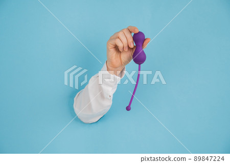 Female hand sticking out of a hole in a blue background and holding a kegel machine.  89847224