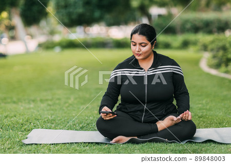 A happy young girl of Indian nationality is sitting in a lotus position in the park with a phone in her hand, resting after a yoga class 89848003