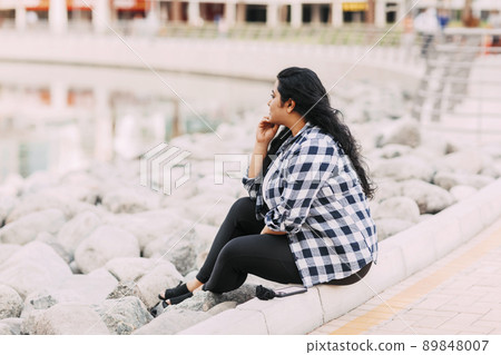 Portrait of a beautiful Indian girl sitting on the embankment and looking thoughtfully into the distance Portrait of a beautiful Indian girl sitting on the embankment and looking thoughtfully into the distance 89848007