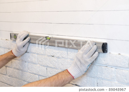 A professional builder sticks decorative tiles on the wall. Uses building level. Flat wall. A worker mounts a decorative brick on a wall 89848967