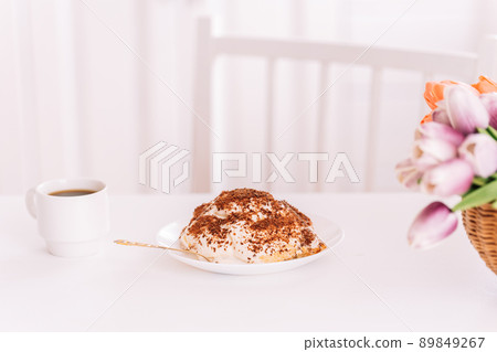 Piece of cake on white plate next to fork and cup of coffee on white table with flowers 89849267
